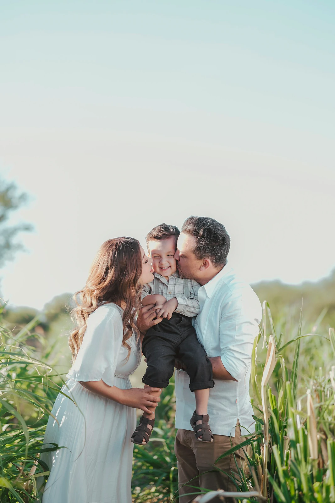 Parents in coordinated white and neutral outfits lifting their toddler in a field of tall grass